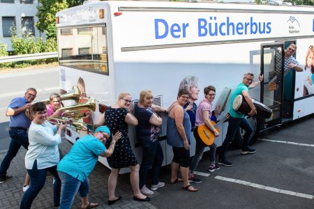 zu sehen sind die  Mitarbeiter der Kreismusikschule mit Instrumenten unter dem Arm, die den Bücherbus anschieben